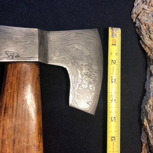 May include: A hand-forged axe with a wooden handle and a patterned steel blade. The blade is approximately 6 inches long.