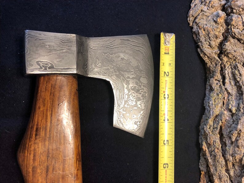 May include: A hand-forged axe with a wooden handle and a patterned steel blade. The blade is approximately 6 inches long.