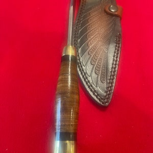 May include: A brown leather knife sheath with a wing design. The sheath has a brass snap closure. The knife has a brown leather handle with a brass pommel.