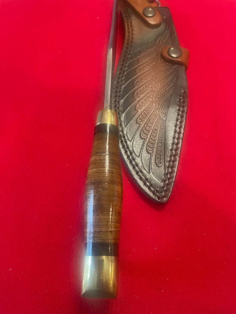 May include: A brown leather knife sheath with a wing design. The sheath has a brass snap closure. The knife has a brown leather handle with a brass pommel.