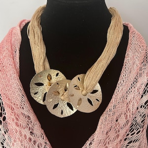 May include: A necklace featuring three silver-colored, sand dollar pendants. The pendants are attached to a beige, multi-strand cord. The necklace is displayed on a black mannequin, with a pink, open-weave scarf in the background.