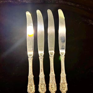 May include: A set of four silver-toned dinner knives with smooth, curved blades and ornate, embossed handles featuring intricate floral and scrollwork detailing. Each knife blade is stamped with the Wallace brand name and sterling silver markings.