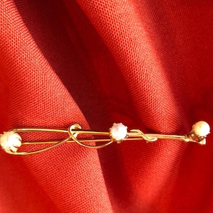 May include: A gold-toned bar pin with three small, round pearls. The pin has a decorative, curved design and is set against a red fabric background. The pin is likely vintage or antique.