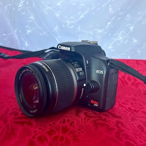 Könnte beinhalten: Eine schwarze Canon EOS Rebel XS digitale Spiegelreflexkamera mit Zoomobjektiv. Die Kamera hat einen schwarzen Riemen und befindet sich vor einem roten Hintergrund. Das Objektiv ist mit "Canon Zoom Lens EF-S" beschriftet.