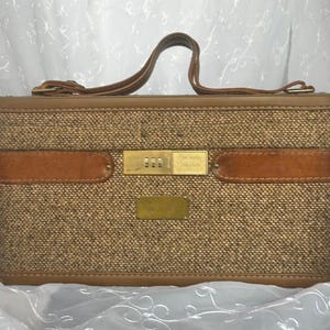 May include: A vintage brown tweed suitcase with leather detailing. The suitcase features a brown leather handle and a combination lock. The rectangular case has a classic, retro design. Likely from the mid-20th century, it's a stylish travel accessory.