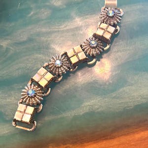 May include: Silver-toned bracelet featuring alternating square and floral designs. The squares have a mother-of-pearl appearance, while the flowers have a central blue gemstone. The bracelet is connected by curved links and has a clasp closure.