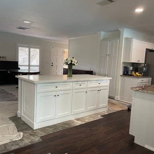 May include: A white kitchen island with a white countertop and four cabinet doors. The island is in a kitchen with white cabinets and a granite countertop.