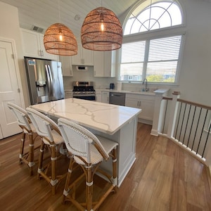 May include: A white kitchen island with a white quartz countertop and three white wicker bar stools. The island is in front of a white kitchen with stainless steel appliances and a large window with white blinds.