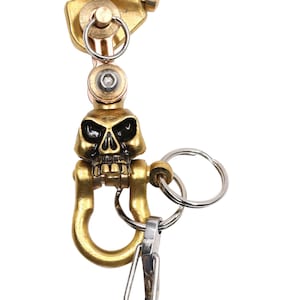 May include: A gold-toned skull-shaped keychain with a brass hook and a smaller silver hook. The keychain also features a smaller brass ring and the text "KYLINK" on the top.