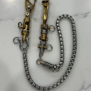 May include: Two metal wallet chains with a skull accent. One chain features a gold-toned swivel clasp and a silver-toned chain. The other chain has a gold-toned skull and a silver-toned chain. Both chains have a swivel clasp.