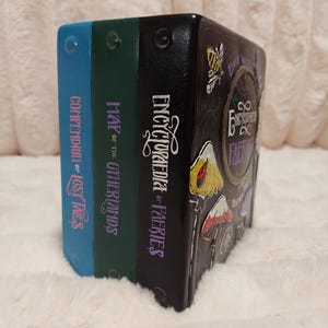 May include: A decorative book set with blue, green, and black spines. The titles include "Compendium of Lost Tales", "Map of the Otherlands", and "Encyclopedia of Faeries". The black book features illustrations of mushrooms, a ladybug, and a bee.