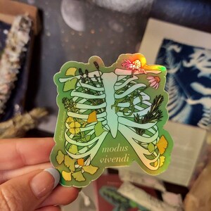 May include: Holographic sticker featuring a rib cage filled with flowers and foliage. The sticker has a green gradient background and the words "modus vivendi" are printed on the lower right. The sticker is held by a person.