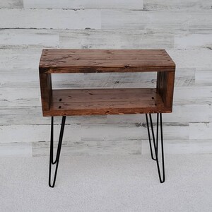 May include: A rustic, wooden side table with a dark stain finish. The table features a rectangular design with an open shelf and is supported by black hairpin legs. The table measures approximately 30 inches long, 12 inches wide, and 20 inches tall.