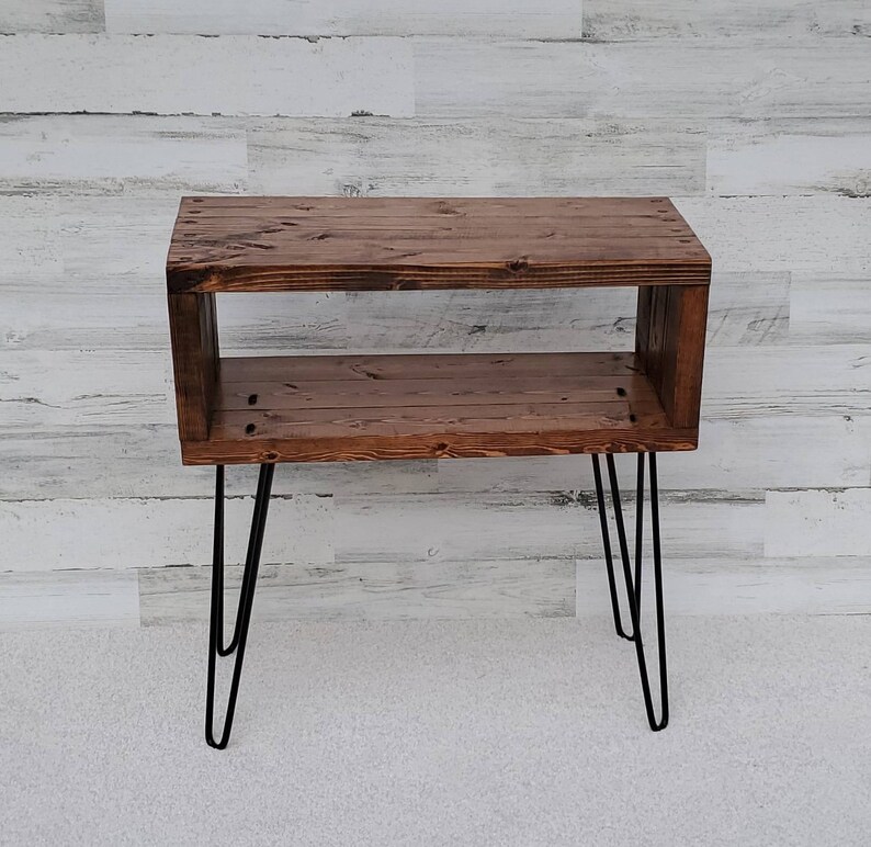 May include: A rustic, wooden side table with a dark stain finish. The table features a rectangular design with an open shelf and is supported by black hairpin legs. The table measures approximately 30 inches long, 12 inches wide, and 20 inches tall.
