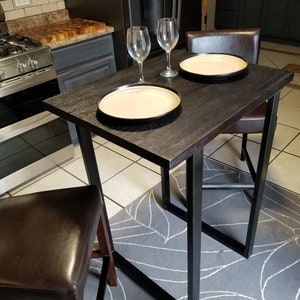May include: A black metal bar table with a dark wood top. Two wine glasses and two plates are on the table. The table is set for two people. The table is in a kitchen with a black refrigerator and a brown leather bar stool.