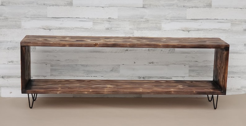 May include: A rustic wooden shelf unit with a dark brown finish. The shelf has a single open shelf and sits on four hairpin legs.
