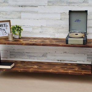 May include: A rustic wooden media console with two shelves, black metal hairpin legs, and a record player on the top shelf. The console has a distressed wood finish and is decorated with a framed sign that says "Rustic Industrial" and a small potted plant.