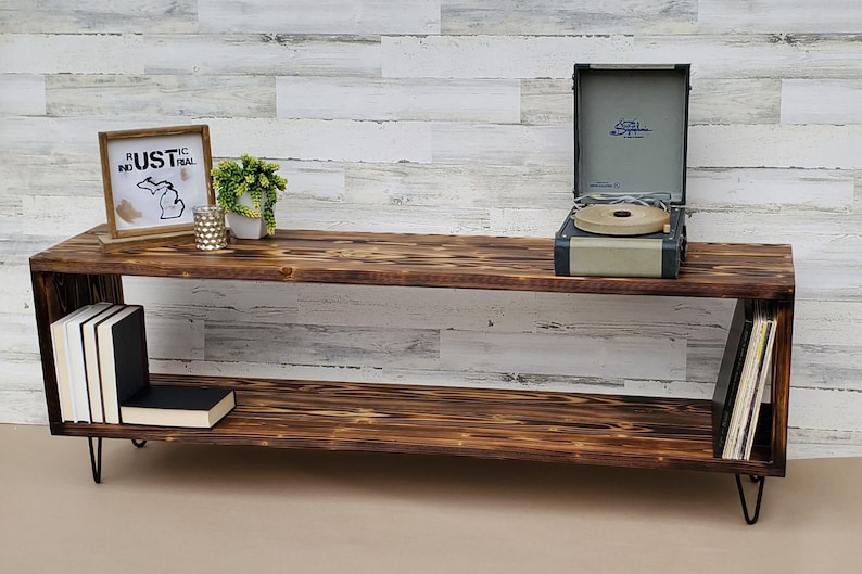 May include: A rustic wooden media console with two shelves, black metal hairpin legs, and a record player on the top shelf. The console has a distressed wood finish and is decorated with a framed sign that says "Rustic Industrial" and a small potted plant.