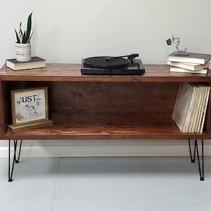 May include: A dark brown wooden record player stand with a shelf for vinyl records. The stand has black hairpin legs and a small shelf on top for a record player. There is a plant in a white pot on the top shelf, and a stack of books on the right side of the stand.