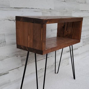 May include: A rustic, wooden side table with a dark stain. The table features a rectangular open box design supported by four black hairpin legs. The wood grain is visible, adding to the natural aesthetic. The table is approximately 24 inches tall.