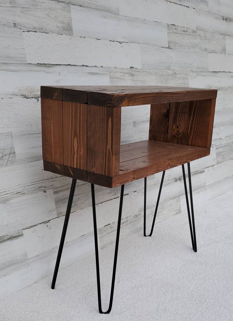 May include: A rustic, wooden side table with a dark stain. The table features a rectangular open box design supported by four black hairpin legs. The wood grain is visible, adding to the natural aesthetic. The table is approximately 24 inches tall.