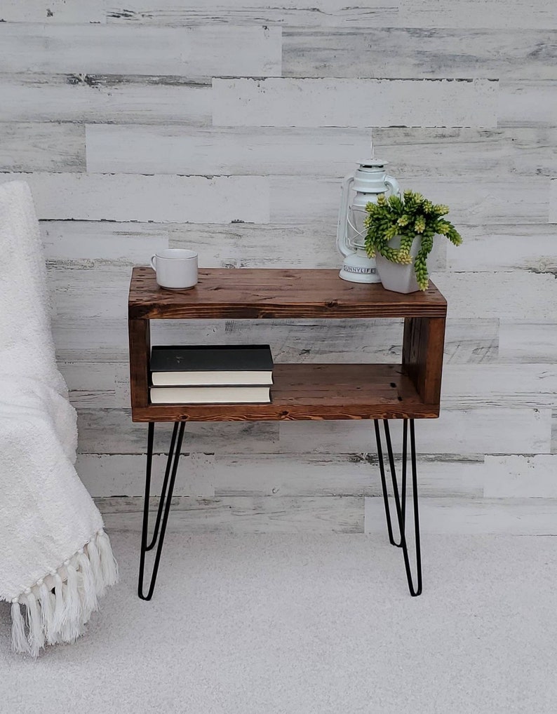 May include: A rustic, brown wooden side table with a lower shelf and black hairpin legs. A white mug, a white lantern, and a small potted plant sit on top. Two books are on the shelf. The table is 24 inches long, 12 inches wide, and 24 inches high.