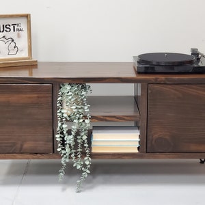May include: A dark brown wooden media console with two doors on each side, a shelf in the middle, and black hairpin legs. The console has a record player on top and a potted plant hanging over the shelf. The console has a sign on top that says "RUSTIC INDUSTRIAL".