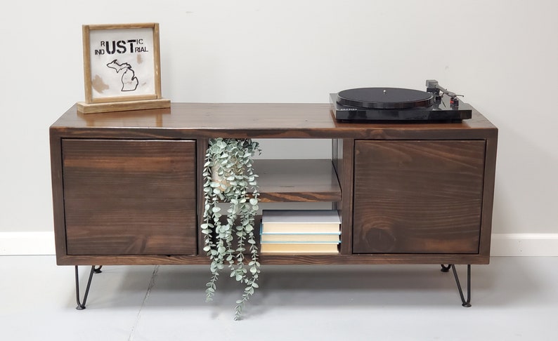May include: A dark brown wooden media console with two doors on each side, a shelf in the middle, and black hairpin legs. The console has a record player on top and a potted plant hanging over the shelf. The console has a sign on top that says "RUSTIC INDUSTRIAL".