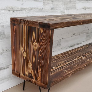 May include: A rustic wooden shelf unit with two shelves and black hairpin legs. The wood has a dark brown, burnt finish.