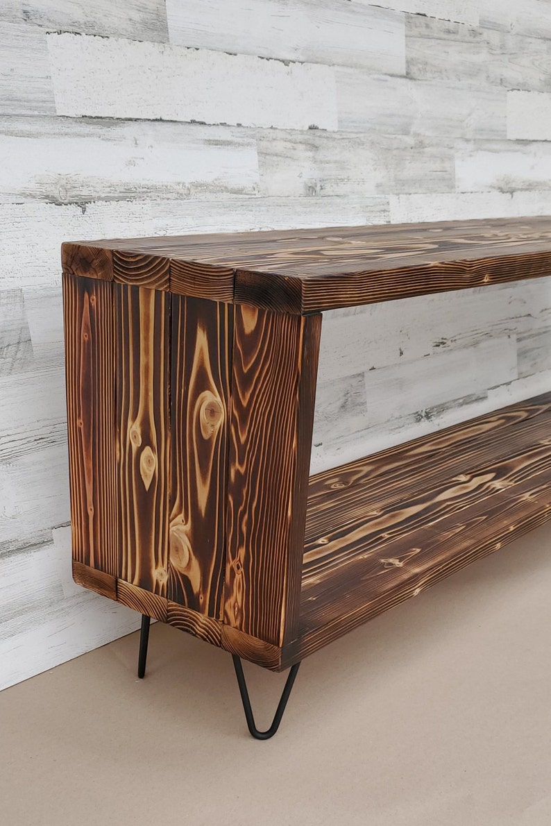May include: A rustic wooden shelf unit with two shelves and black hairpin legs. The wood has a dark brown, burnt finish.