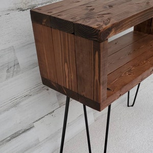 May include: A handcrafted wooden side table with a rich brown stain. The table features a rectangular design with an open shelf and is supported by black hairpin legs. The wood grain is visible, adding to its rustic charm.