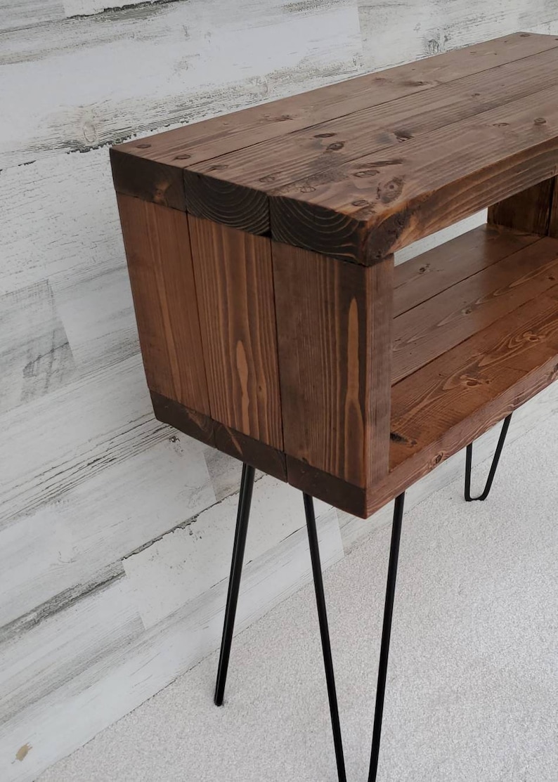 May include: A handcrafted wooden side table with a rich brown stain. The table features a rectangular design with an open shelf and is supported by black hairpin legs. The wood grain is visible, adding to its rustic charm.
