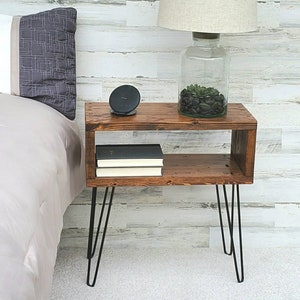 May include: A rustic, wooden nightstand with a warm brown finish. It features a lower shelf and is supported by black hairpin legs. A lamp with a beige shade and a glass base sits on top, along with a wireless charger and decorative items.
