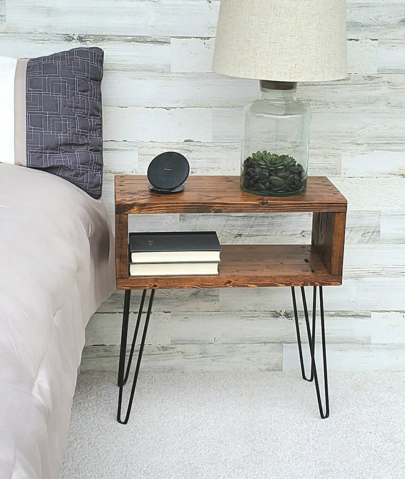 May include: A rustic, wooden nightstand with a warm brown finish. It features a lower shelf and is supported by black hairpin legs. A lamp with a beige shade and a glass base sits on top, along with a wireless charger and decorative items.