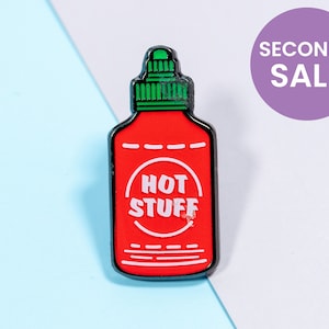 Pode incluir: Pin de esmalte vermelho e preto em forma de garrafa de molho picante com as palavras "HOT STUFF" em um círculo branco na frente.
