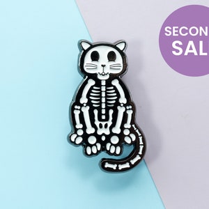 Puede incluir: Pin de esmalte negro con un gato esqueleto con huesos blancos. El gato está sentado con la cola enroscada alrededor de su cuerpo. El pin tiene un acabado brillante.