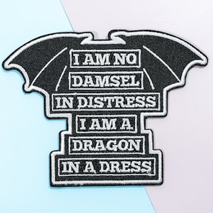 May include: Black embroidered iron-on patch with white text that reads "I am no damsel in distress I am a dragon in a dress". The patch features a black dragon wing design.