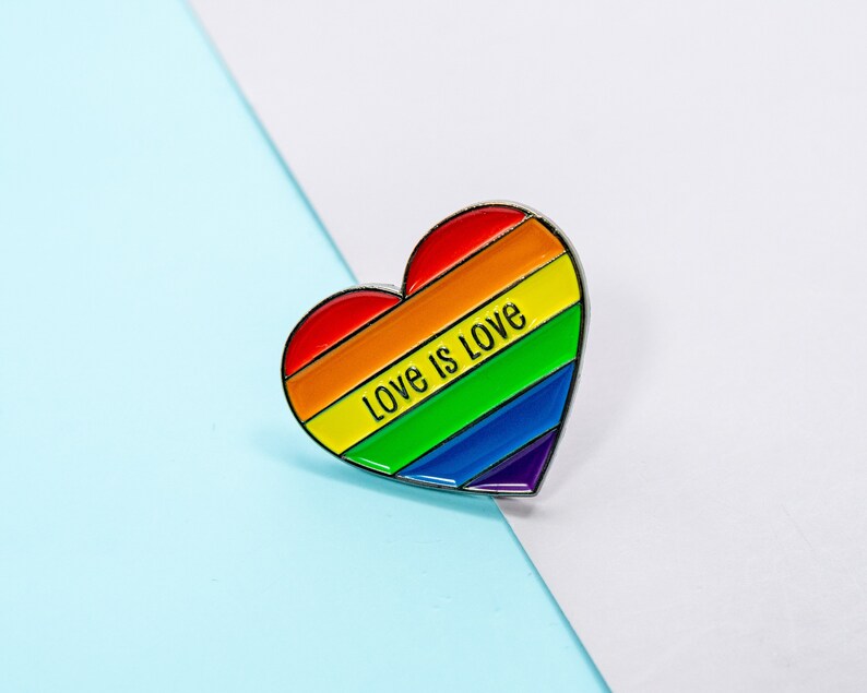 Op de afbeelding: Een regenboogvormige hartvormige pin met de tekst "Love is Love" in zwarte letters.