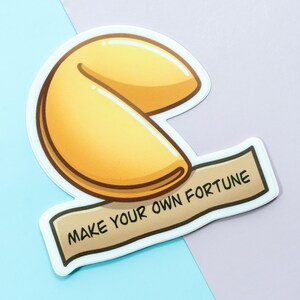 May include: A white sticker with a cartoon illustration of a fortune cookie. The cookie is yellow and brown and has the text "MAKE YOUR OWN FORTUNE" on a brown banner.