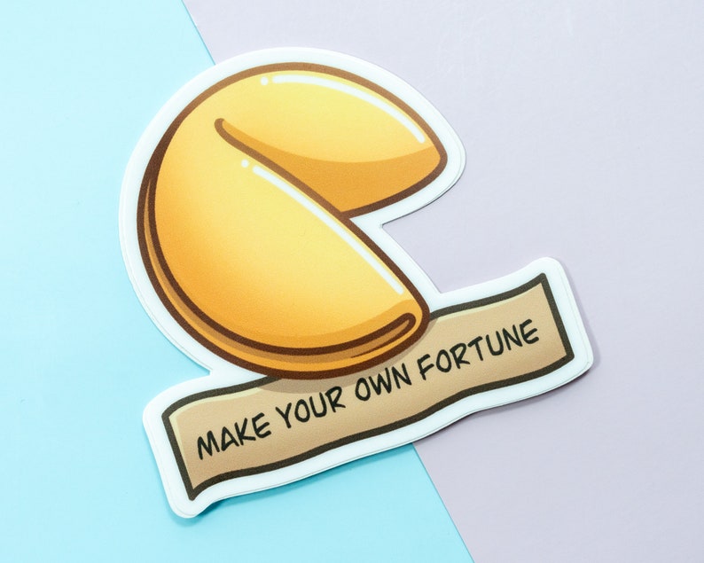 May include: A white sticker with a cartoon illustration of a fortune cookie. The cookie is yellow and brown and has the text "MAKE YOUR OWN FORTUNE" on a brown banner.