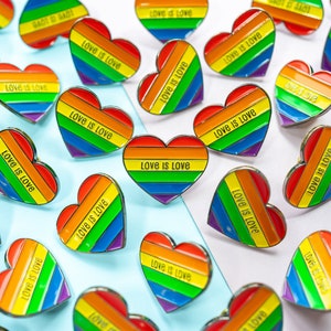 Op de afbeelding: Een verzameling regenboogvormige hartvormige pinnen met de tekst "Love is Love" op elke pin. De pinnen zijn gerangschikt op een lichtblauw en wit oppervlak.