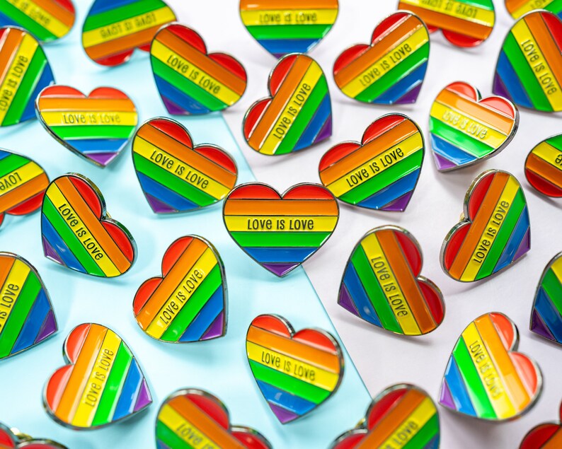 Op de afbeelding: Een verzameling regenboogvormige hartvormige pinnen met de tekst "Love is Love" op elke pin. De pinnen zijn gerangschikt op een lichtblauw en wit oppervlak.