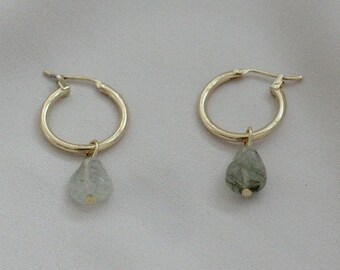 May include: A pair of gold hoop earrings with a small, teardrop-shaped stone dangling from each hoop. The stone on the left earring is clear, while the stone on the right earring is a dark green.