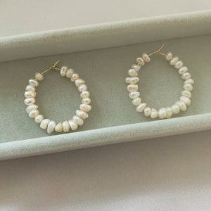 May include: A pair of hoop earrings featuring small, irregular white freshwater pearls strung along a thin gold-toned metal wire. The circular earrings have a simple clasp mechanism at the top, showcasing the natural texture and lustre of the pearls.