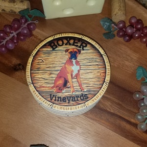 May include: A set of four brown and yellow coasters with a wood grain design. The coasters feature a brown and white Boxer dog sitting on a wooden barrel with the text "Boxer Vineyards".