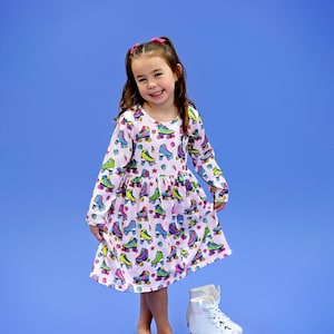 May include: A young girl wearing a pink dress with a roller skate pattern. She is smiling and wearing pink hair ties. She is standing on a blue background and wearing white roller skates with pink wheels.