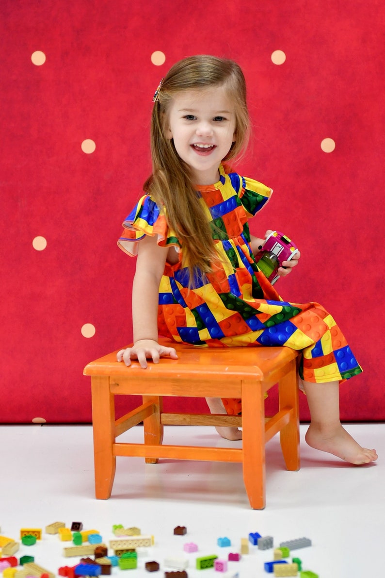 May include: A young girl wearing a colourful dress with a Lego pattern sits on a wooden stool. She is holding a small toy car in her hand. There are colourful Lego blocks scattered on the floor in front of her.