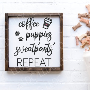 May include: A wooden sign with the text "coffee, puppies, sweatpants, repeat" in black lettering. The sign is framed in brown wood and has a white background. A white ceramic jar with the text "good dog" in black lettering is in the background.