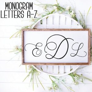 May include: A wooden sign with a white background and a brown frame. The sign features a cursive monogram of the letters "E", "D", and "L". The sign is surrounded by white flowers and green leaves.