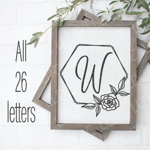 May include: A wooden sign with a white background and a black hexagonal frame. The letter "W" is inside the hexagon with a black floral design. The sign is on a white brick wall with green branches.
